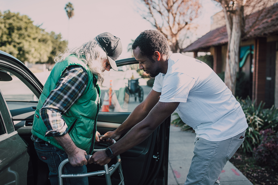 A younger man helps an elderly man with a walker out of his car